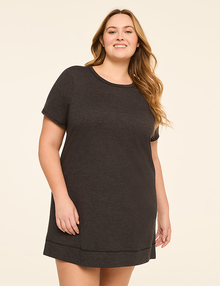 A smiling woman in a dark grey t-shirt dress stands against a plain, light beige background.