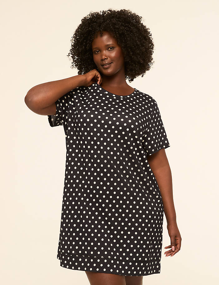 A woman with dark, curly hair stands in a black and white polka dot dress against a plain, light-colored background.