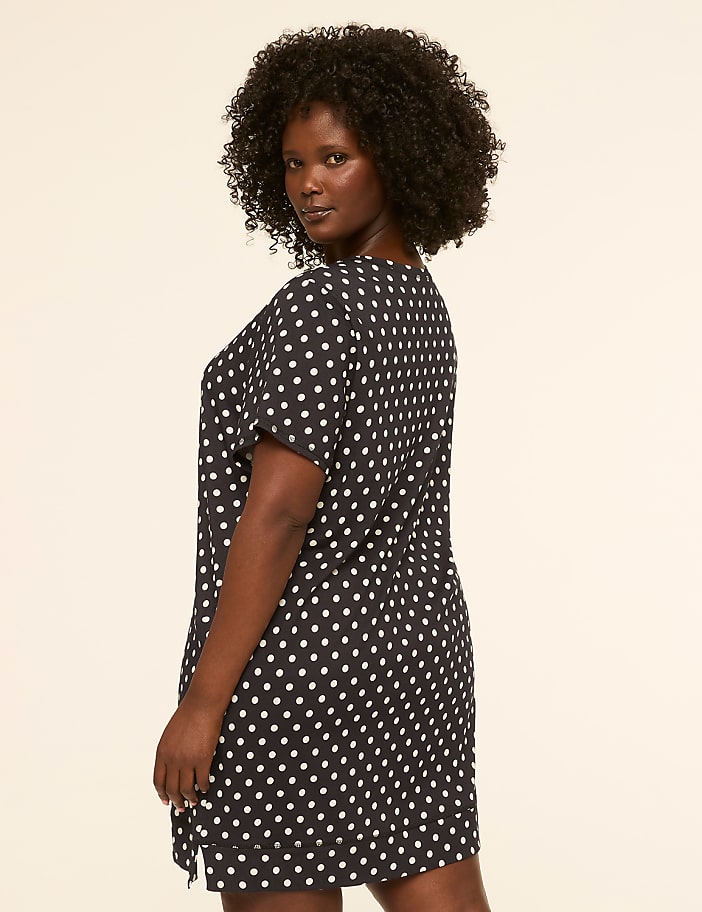 A woman with dark, curly hair poses in a dark, polka-dotted dress against a plain, light-colored background.