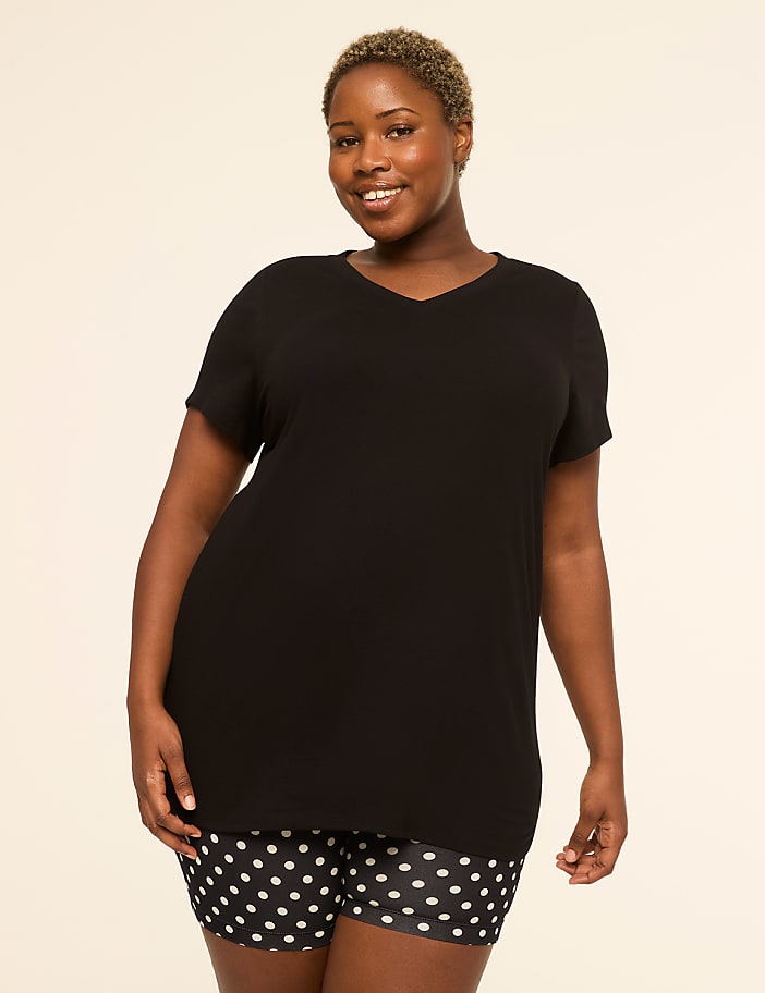 A smiling woman with short blonde hair poses in a black v-neck shirt and polka dot shorts against a plain, light background.