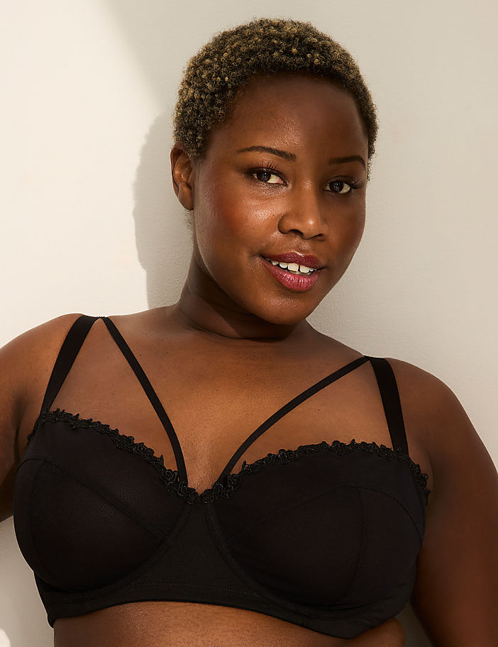 A woman with short, curly hair smiles at the camera, wearing a black bra with strappy detailing, against a plain white background.