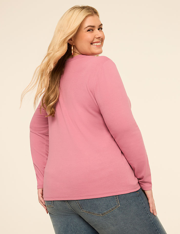 A woman with blonde hair wearing a pink long-sleeved shirt and blue jeans smiles over her shoulder against a plain background.