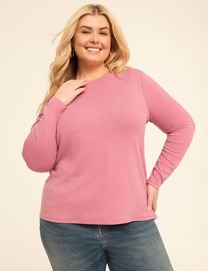 A smiling woman in a pink long-sleeved shirt and blue jeans stands against a plain, light-colored background.
