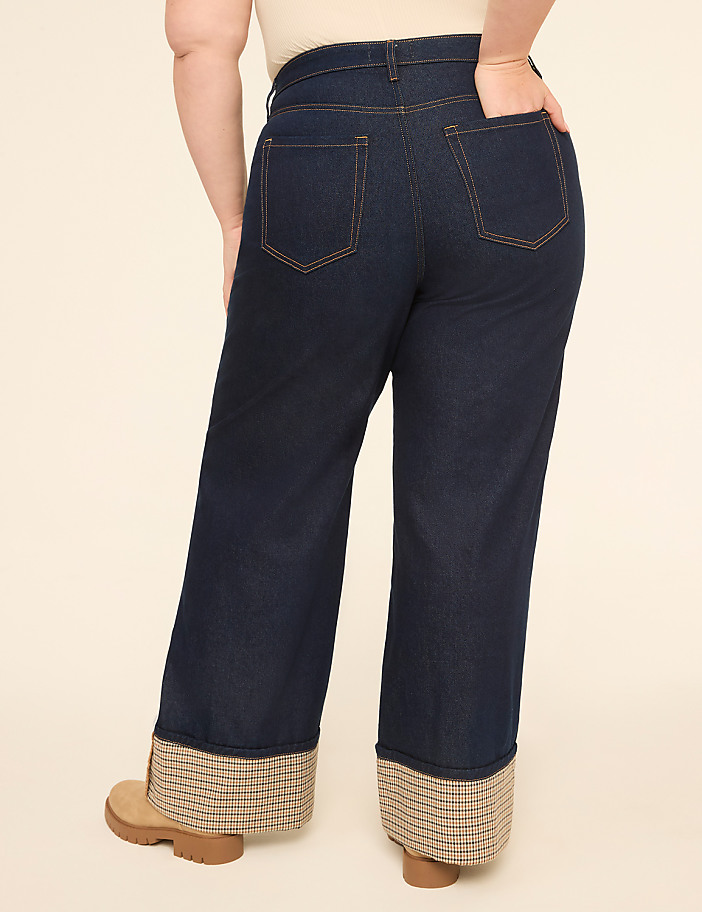 A person wearing dark wash denim jeans with cuffed hems stands against a plain, light-colored background.