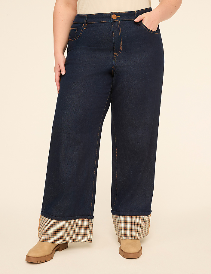 A person stands in dark wash wide-leg jeans with plaid cuffs and tan boots against a plain, light-colored background.