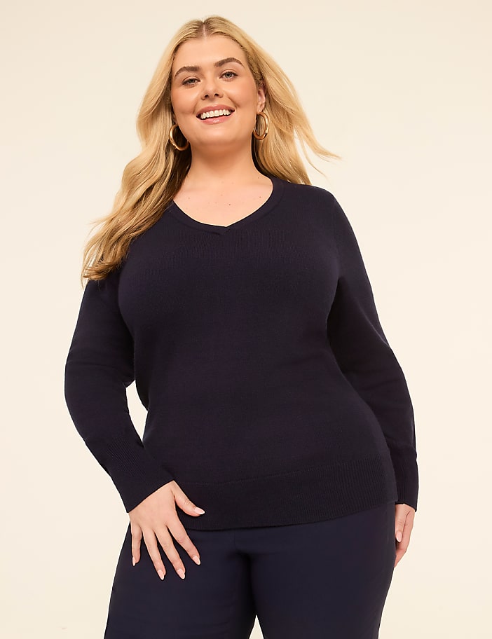A smiling woman in a navy v-neck sweater and dark pants stands against a plain, light-colored background.