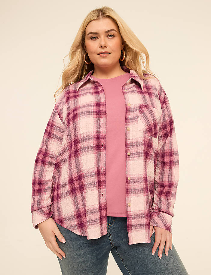 A woman in a pink plaid shirt and jeans stands against a plain, light-colored background.