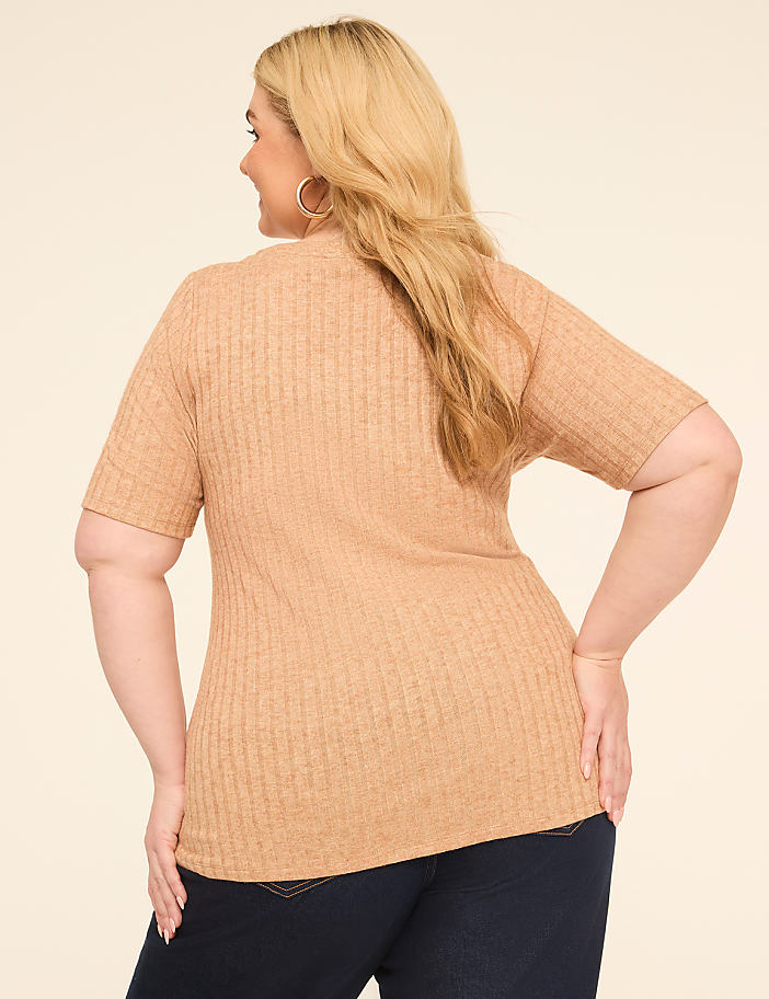 A woman in a tan ribbed top and dark jeans stands against a plain cream background.