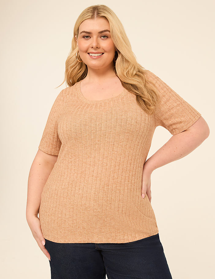 A smiling woman with blonde hair wears a tan ribbed shirt and dark jeans against a plain beige background.