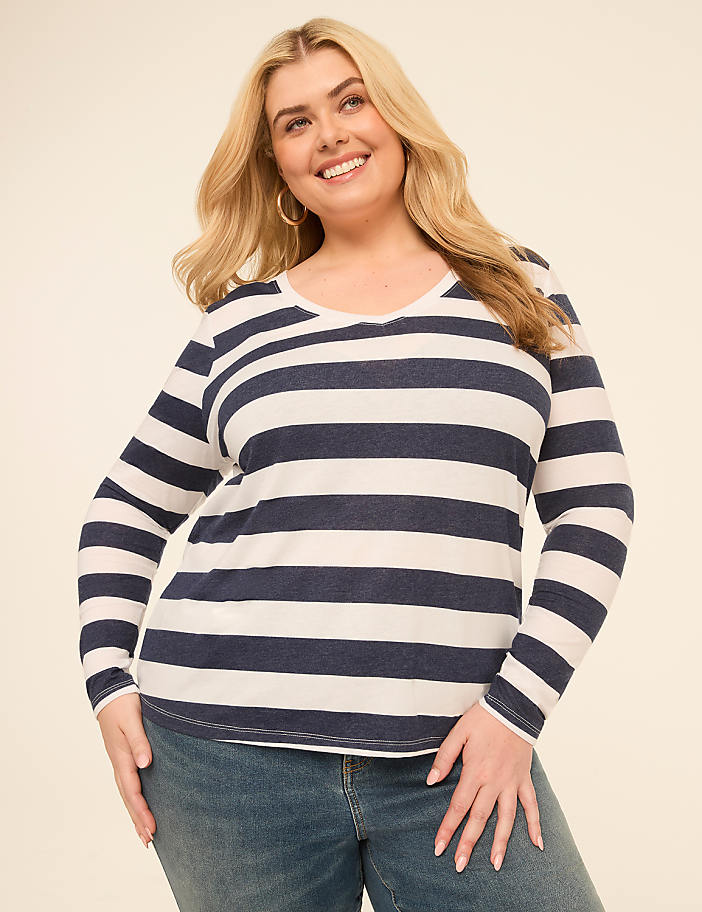 A smiling woman with blonde hair wears a striped long-sleeved shirt and jeans against a plain, light-colored background.