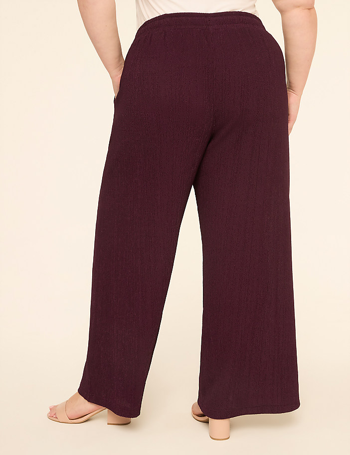 A person wearing burgundy ribbed wide-leg pants stands with their back to the camera against a plain cream background.