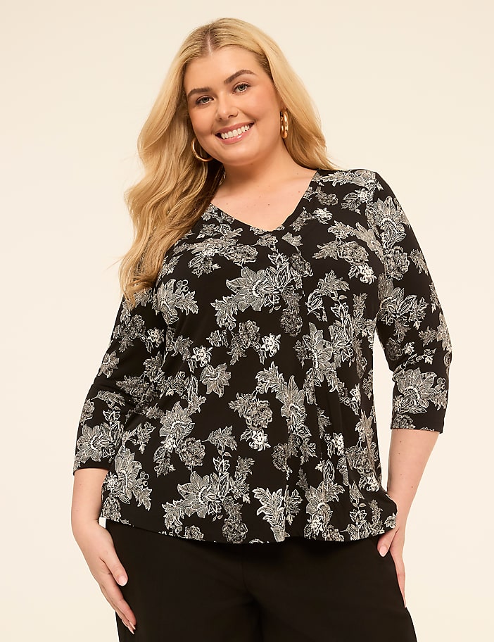 A smiling woman with blonde hair and a black and white floral top stands against a plain, light-colored background.