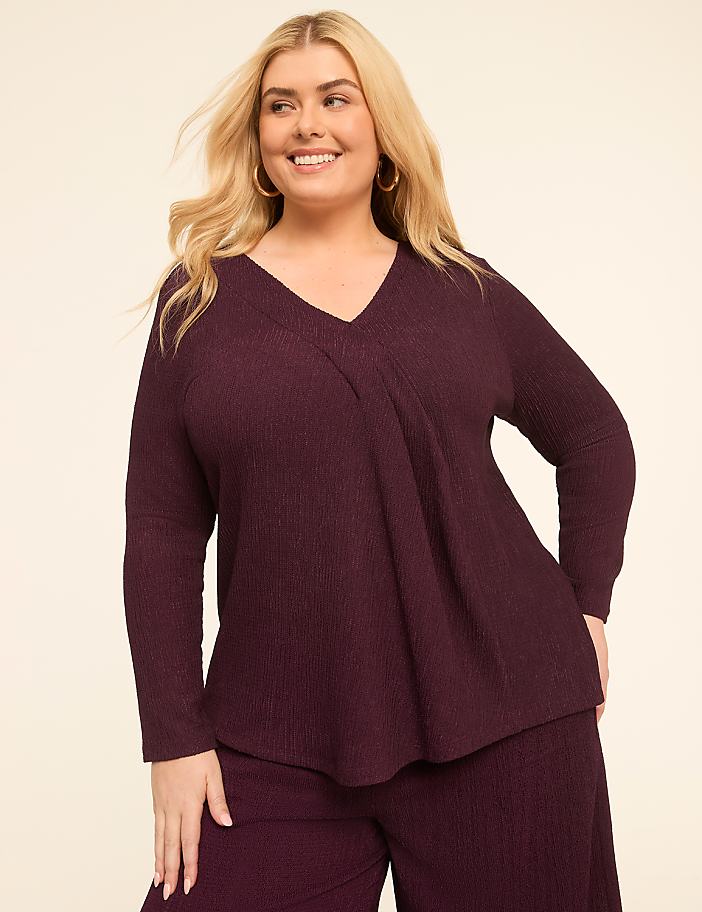 A smiling woman in a plum-colored ribbed top and pants stands against a plain, light-colored background.