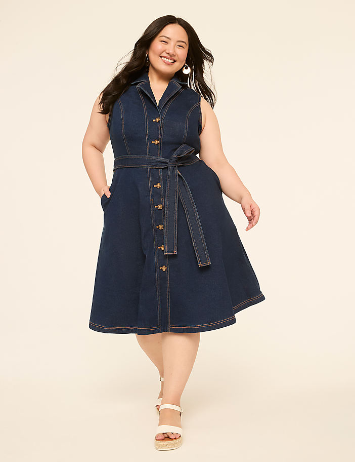A smiling woman in a denim dress stands against a plain, light-colored background.