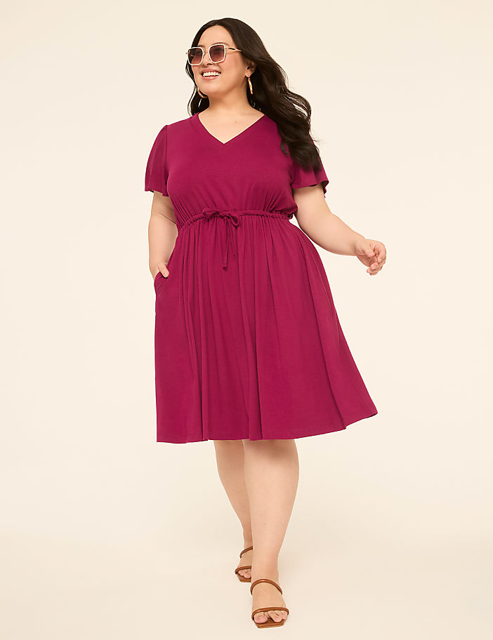 A woman in a magenta dress with pockets and sandals stands against a plain, light-colored background.