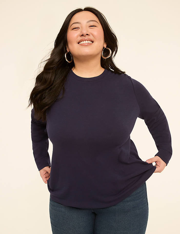 A smiling woman in a navy long-sleeved shirt and jeans stands against a plain, light-colored background.