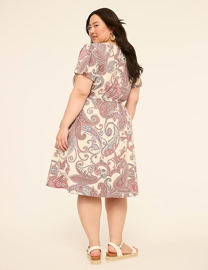A woman in a paisley print dress stands against a plain, light-colored background.