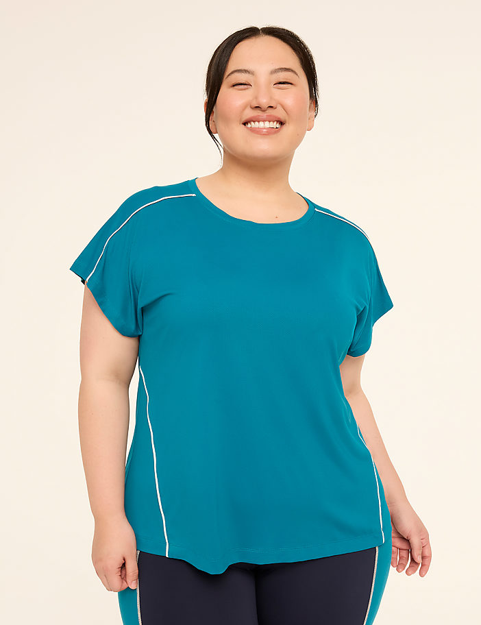 A smiling woman in a teal t-shirt with white piping stands against a plain, light-colored background.