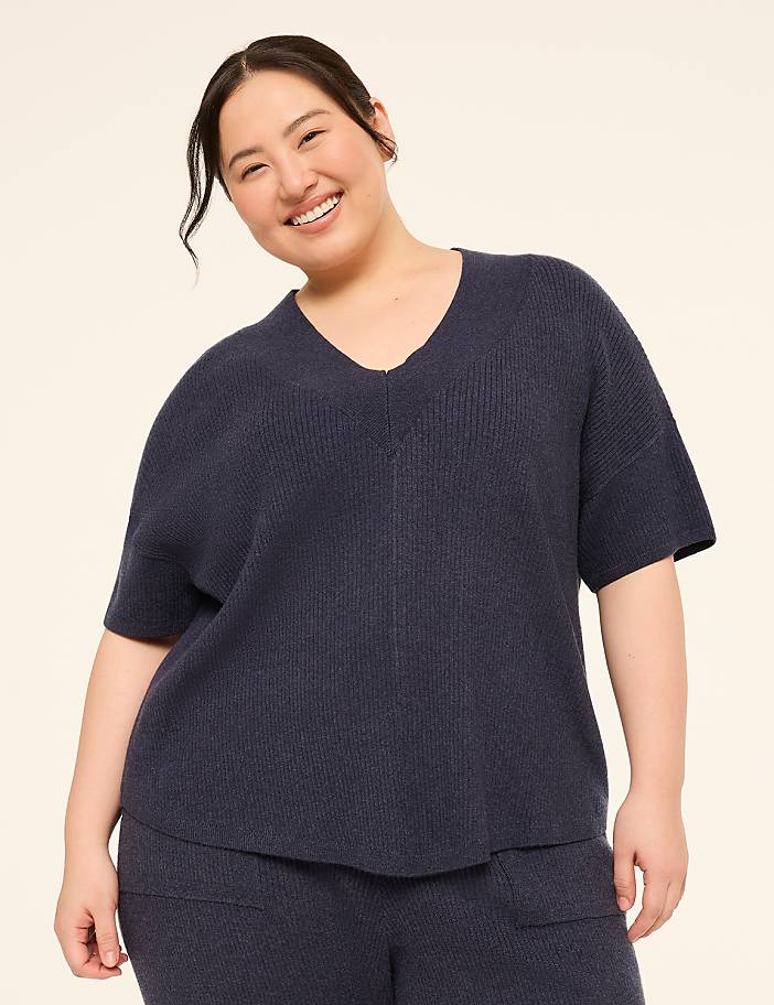 A smiling woman in a navy blue v-neck sweater and matching pants stands against a plain, light beige background.