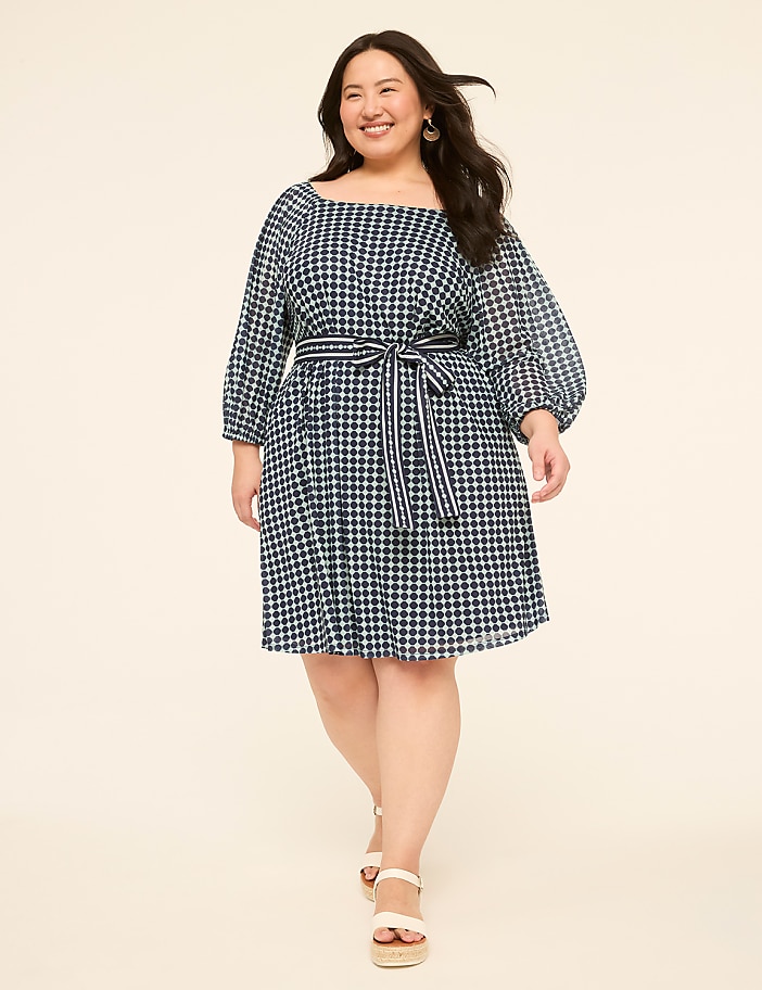 A woman in a blue and white checkered dress with puffed sleeves and a sash walks forward against a plain, light-colored background.