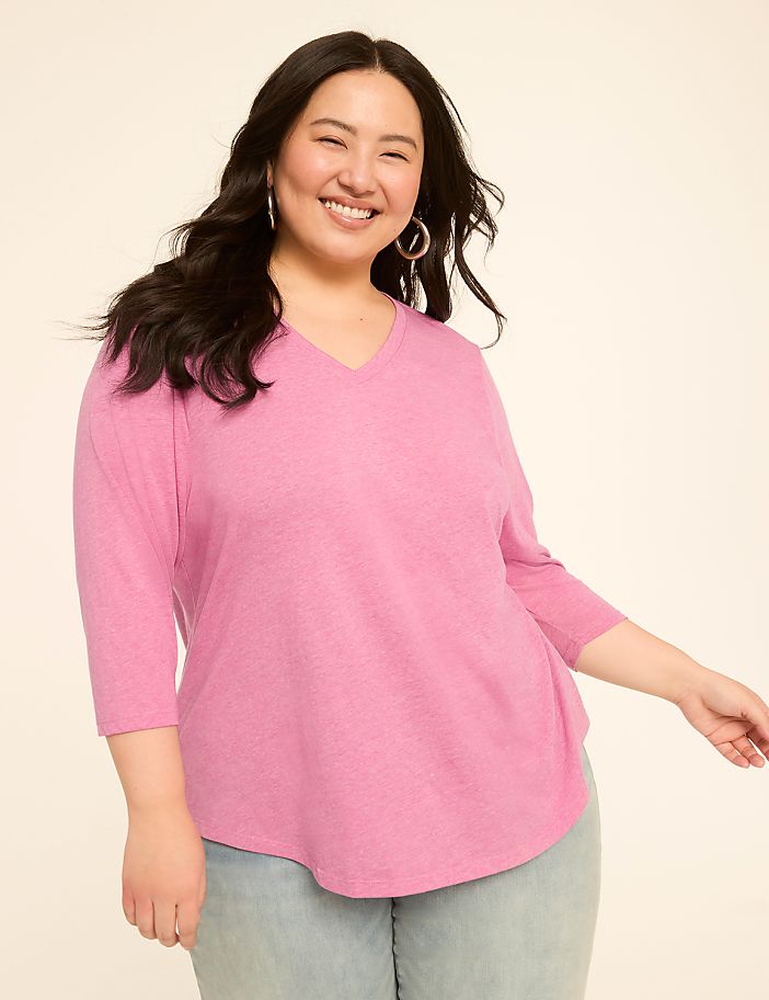 A smiling woman in a pink v-neck shirt and light blue jeans poses against a plain, light-colored background.