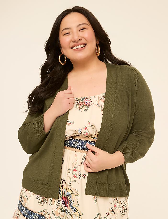 A smiling woman in a green cardigan and floral dress stands against a plain, light-colored background.