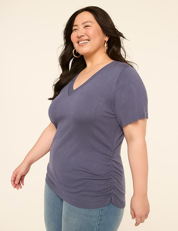 A smiling woman in a blue v-neck shirt and jeans stands against a plain, light-colored background.