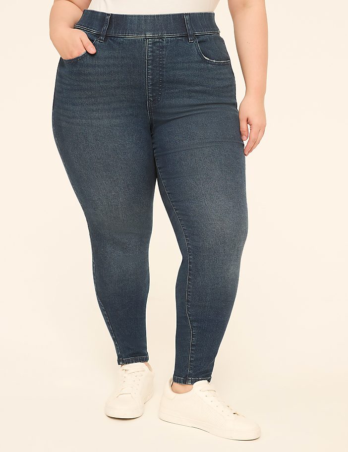 A person in dark wash denim jeans and white sneakers stands against a plain, light-colored background.