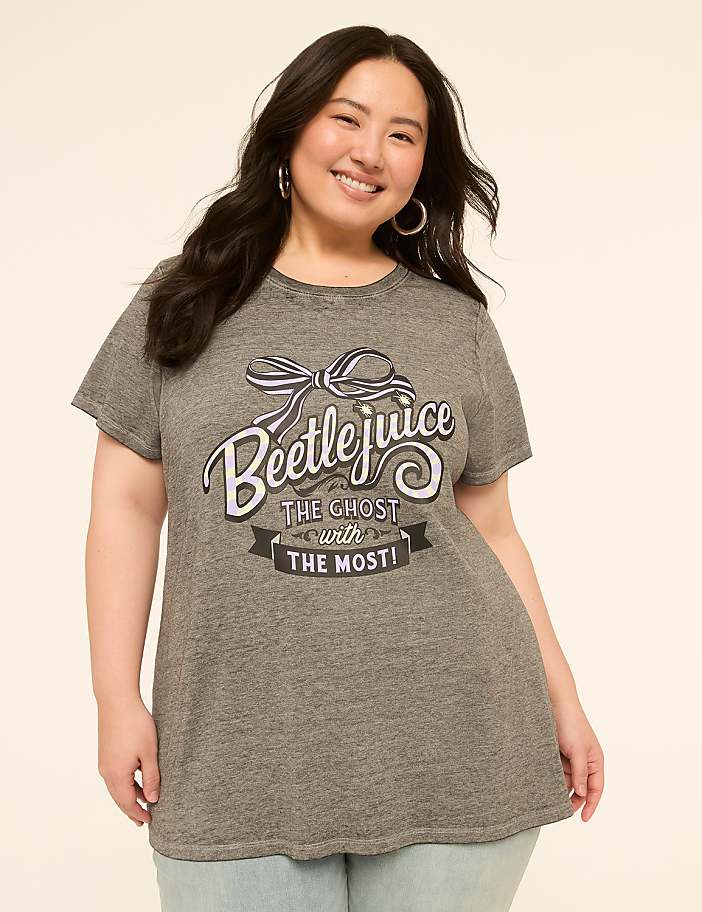A smiling woman in a grey Beetlejuice t-shirt stands against a plain, light-colored background.