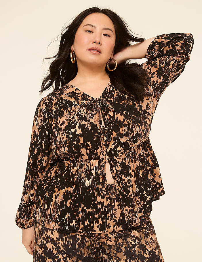 A woman with dark hair and hoop earrings poses in a patterned top and pants against a plain, light-colored background.