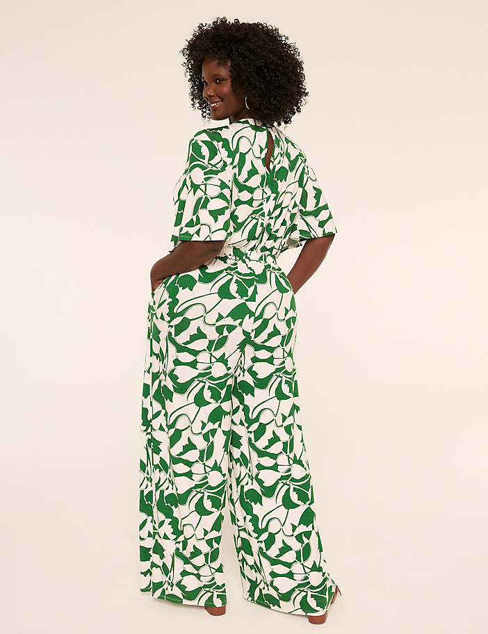 A woman in a green and white patterned jumpsuit stands with her hands in her pockets against a plain, light-colored background.