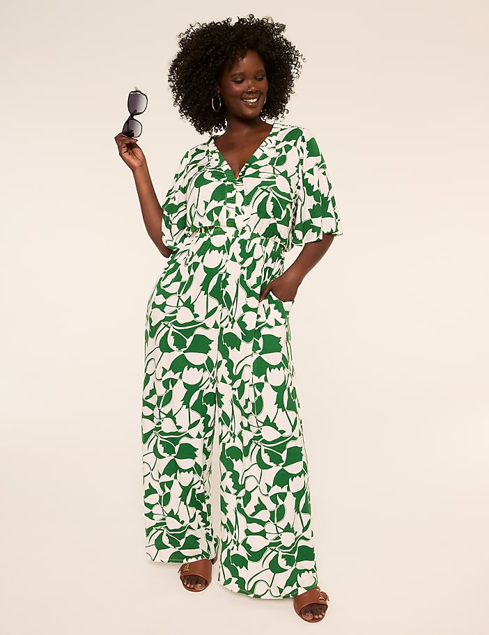 A smiling woman in a green and white floral jumpsuit stands with sunglasses in her hand against a plain, light background.