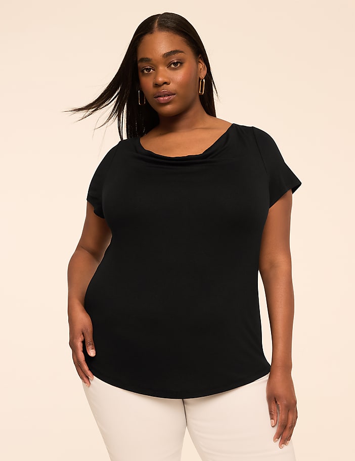 A woman in a dark brown cowl-neck top and white pants stands against a plain, light beige background.