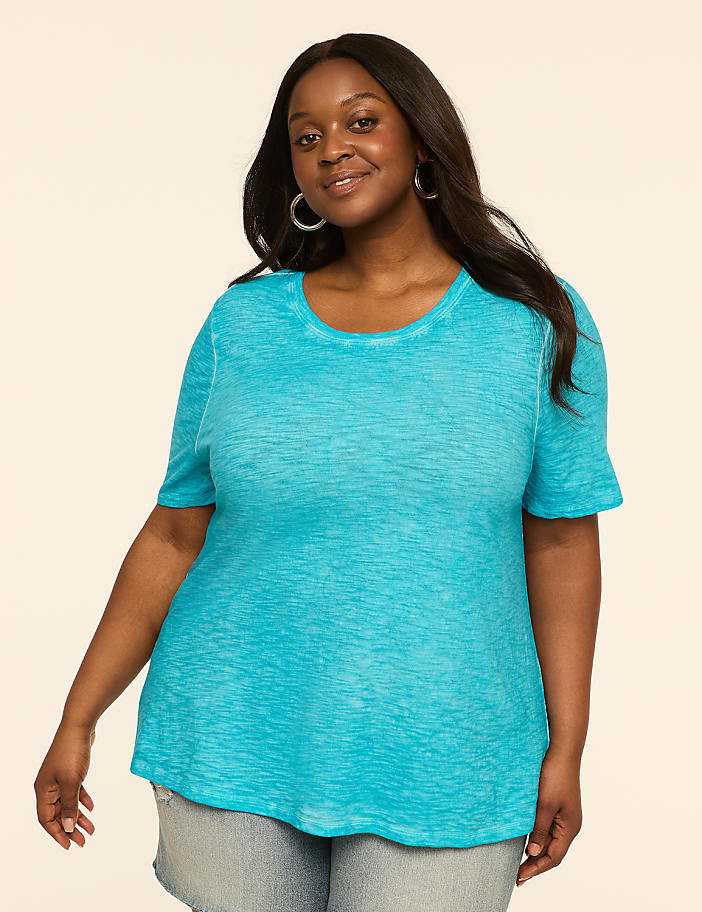 A woman in a turquoise t-shirt and denim shorts smiles against a plain, light background.