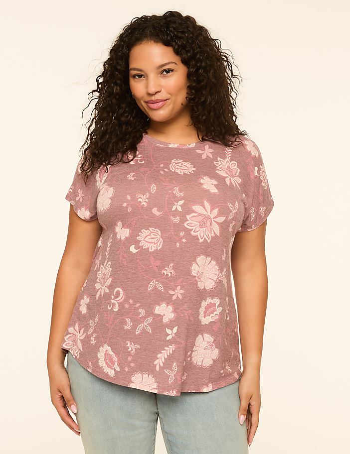 A woman with curly dark hair smiles at the camera, wearing a pink floral t-shirt and light blue jeans against a plain, light-colored background.