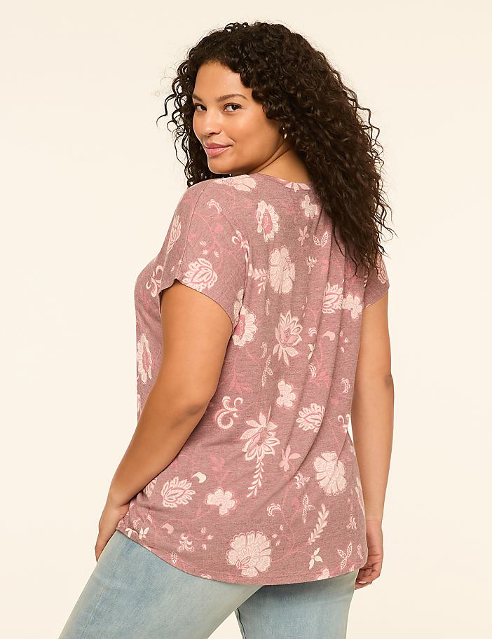 A woman with curly dark hair, wearing a floral print top and light blue jeans, poses against a plain, light-colored background.