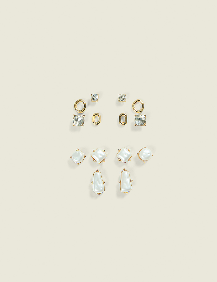 A collection of assorted gold earrings with clear stones and pearls are arranged on a pale cream background.