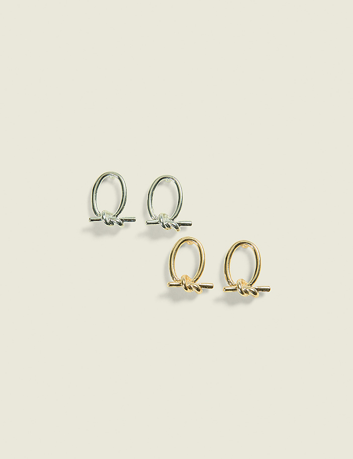 Two pairs of minimalist knot-shaped earrings, one silver and one gold, rest on a soft, pale yellow background.