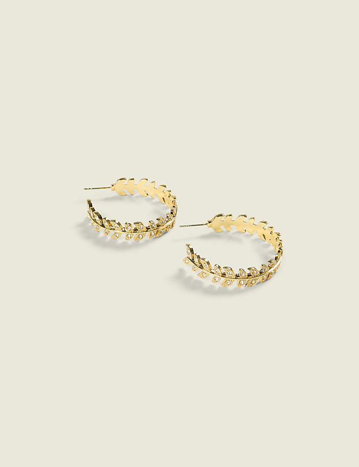 A pair of gold hoop earrings with a leaf design rests on a plain, light-colored background.