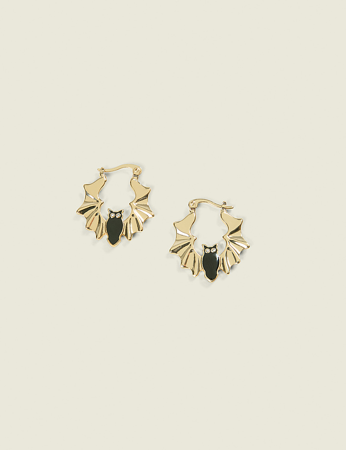 A pair of gold bat-shaped hoop earrings with black enamel details rests on a pale yellow background.