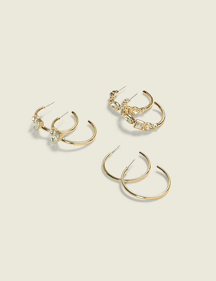 Three pairs of gold hoop earrings, two adorned with clear stones, rest on a pale, textured background.