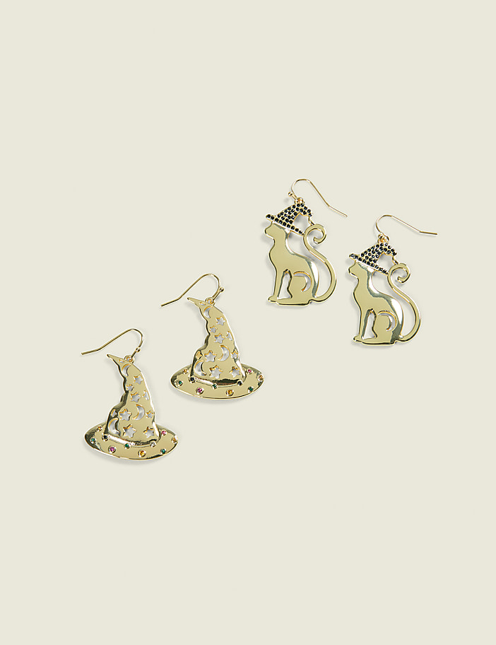 Two pairs of gold-toned Halloween-themed earrings, one featuring witch hats and the other cats wearing witch hats, rest on a plain cream-colored background.