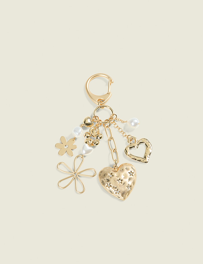 A gold keychain with various charms, including a textured heart, a wire flower, a pearl, and a small flower, hangs against a pale yellow background.