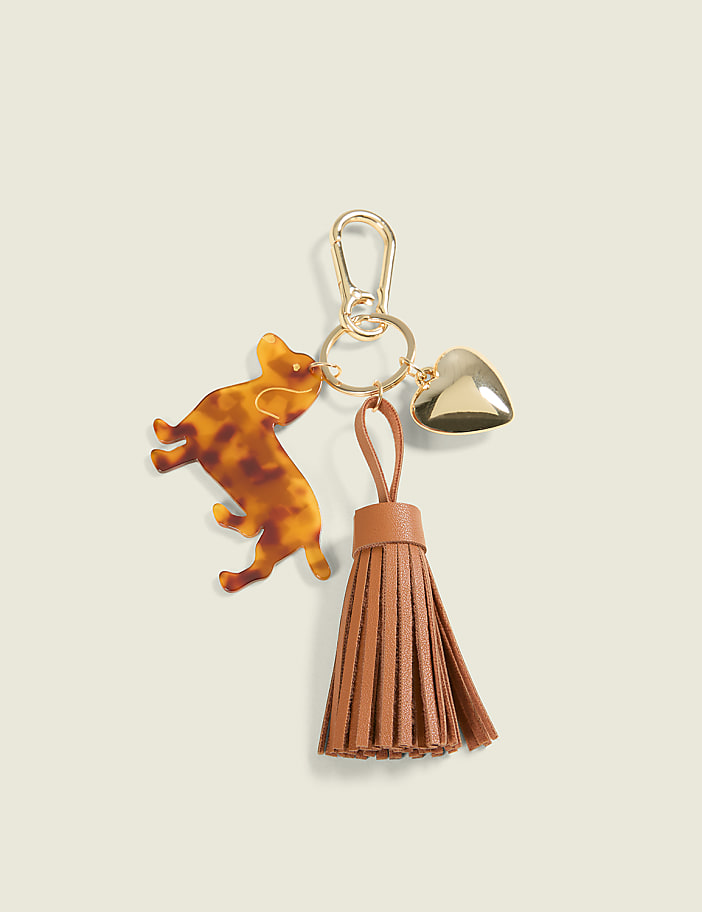 A tortoiseshell cat charm, a gold heart charm, and a brown leather tassel hang from a gold keychain against a plain, light brown background.