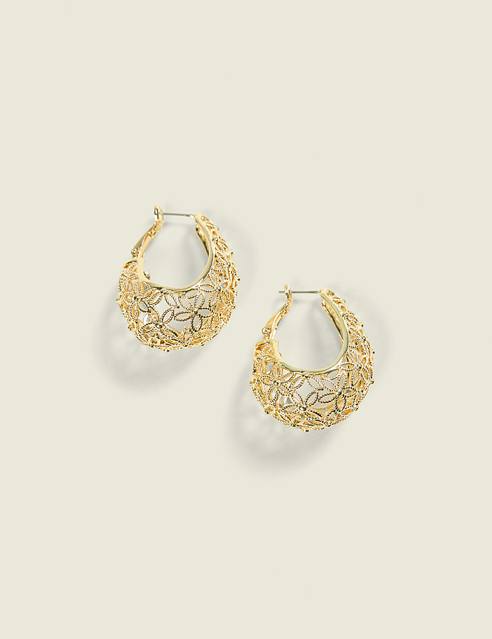 A pair of ornate gold hoop earrings with floral cutouts rests on a pale, neutral background.