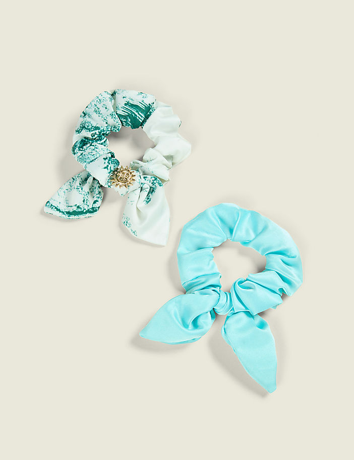 Two scrunchies, one with a marbled green and white pattern and a gold accent, and another in a solid bright blue, rest on a pale yellow background.