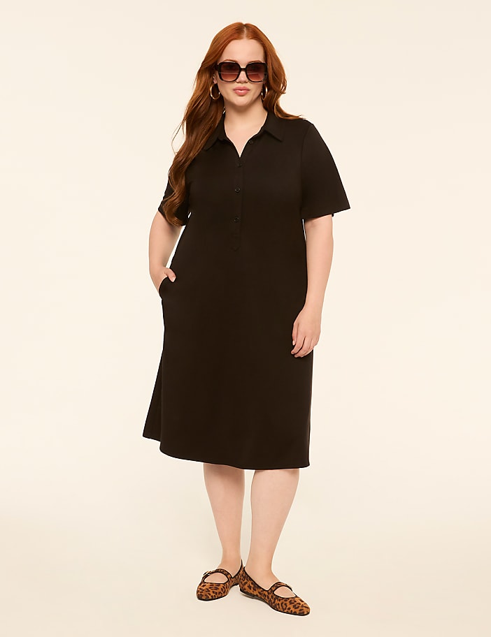 A woman with red hair and sunglasses stands in a black dress and leopard print flats against a plain, light-colored background.