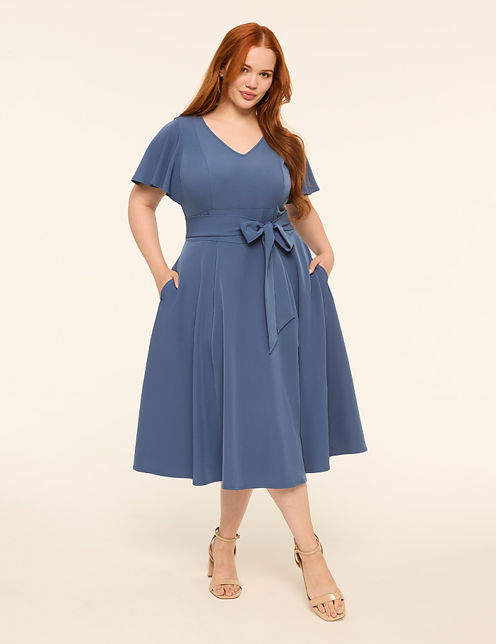A woman in a blue dress stands with her hands in her pockets against a plain, light background.