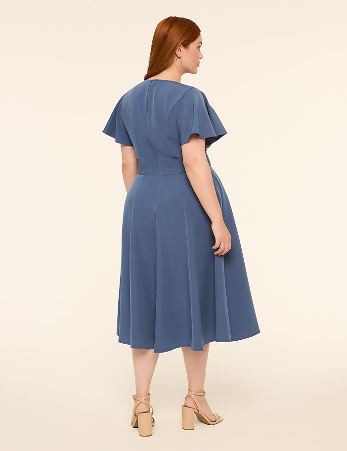 A woman in a blue dress stands against a plain, light-colored background.