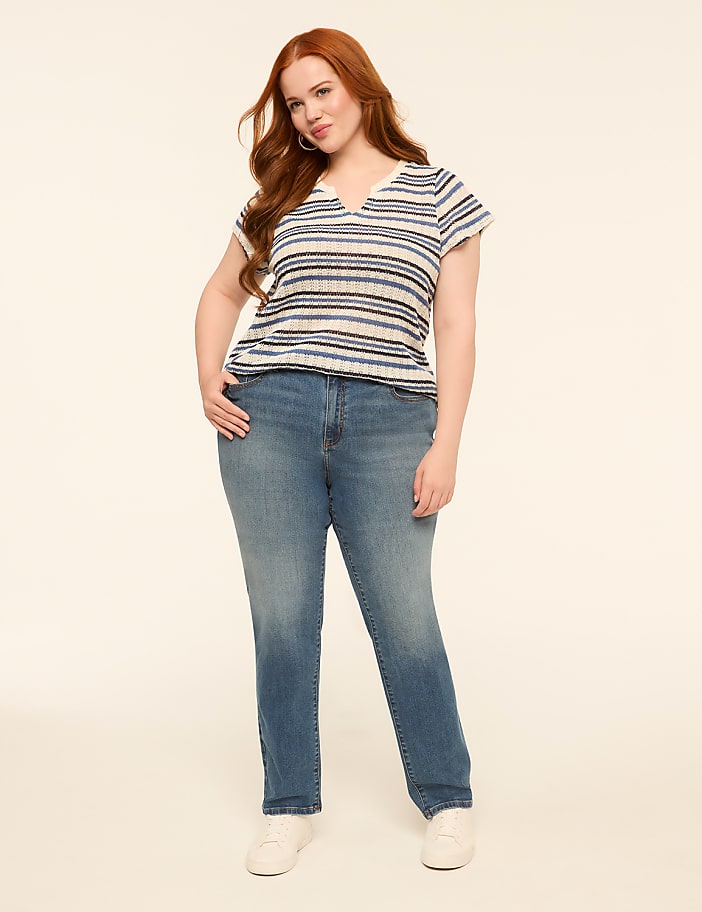 A woman with long red hair stands in the foreground wearing a striped shirt and jeans against a plain, light-colored background.